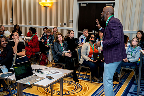 A presenter standing in front of seated conference session attendees.