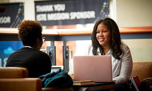 Two Black women seated together in a lounge area. One has her laptop open but they are looking at each other.