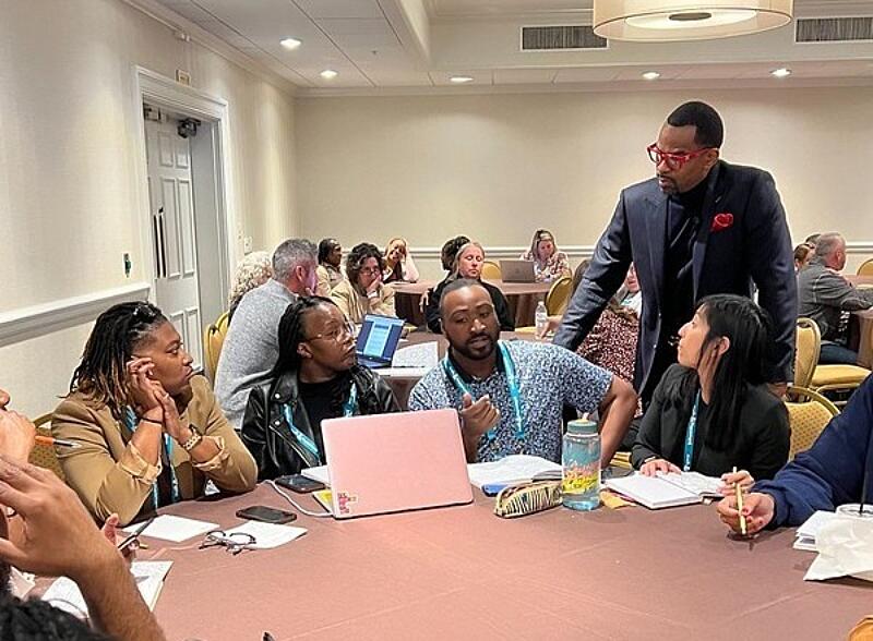 Black male professional in a conference room with diverse adult attendees seated at tables.