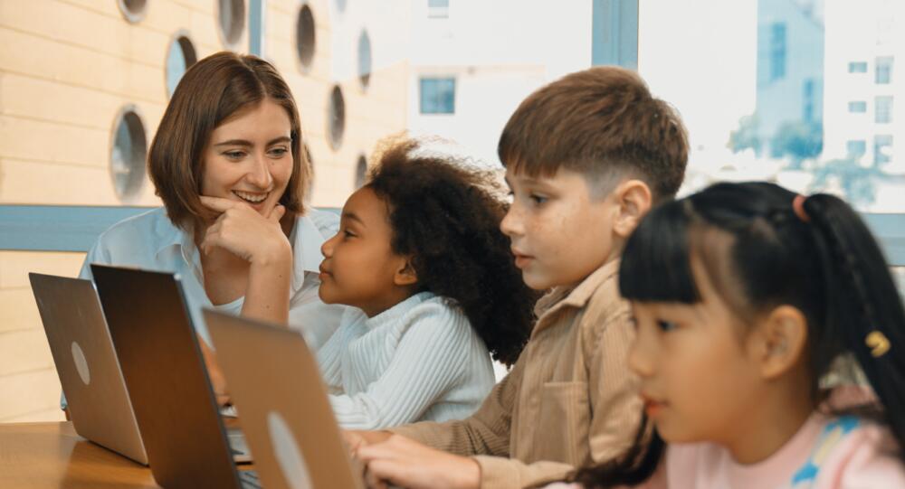 A small group of diverse students seated at a table looking at laptops. A teacher is seated at the end of the table and is smiling at one of the students.
