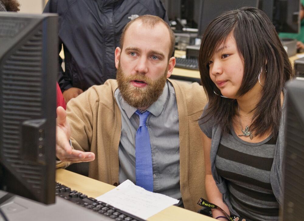 A white male teacher and an Asian female student both looking at a computer monitor in a computer lab classroom.