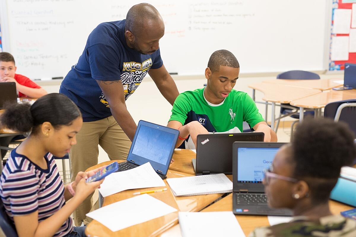 A Black male teacher is observing a Black male student working at a table with other students engaged in schoolwork.