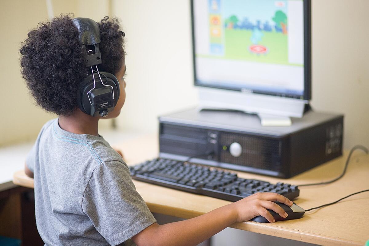 A young Black student seated facing away from the camera wearing headphones and working at a computer station.