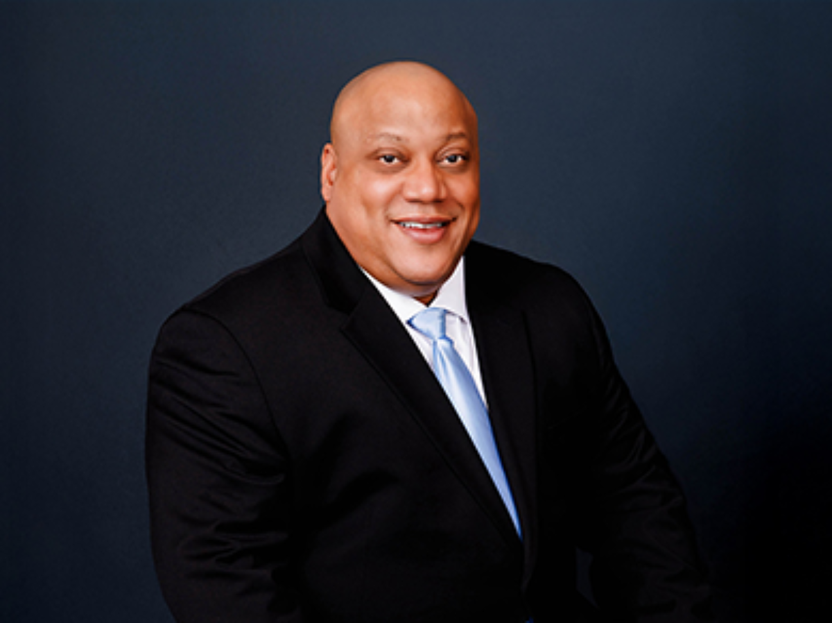 A man in a black suit jacket, white shirt, and light blue tie smiles in front of a dark blue background.