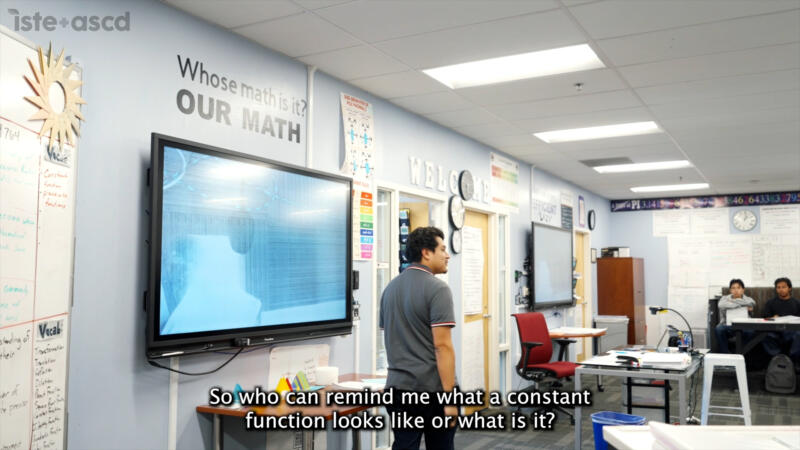 A teacher stands before a digital display in a math classroom with the text So who can remind me what a constant function looks like or what is it? overlaid at the bottom.