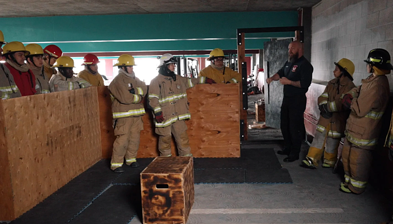 A group of people in firefighting gear stand behind wooden barriers, listening to an instructor in a dark uniform. They are indoors, possibly in a training facility.