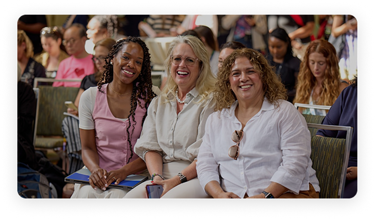 Three women sitting in the front row at an event, smiling and looking toward the camera, with other people seated and talking in the background.