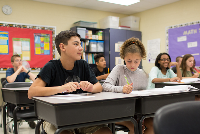Two students of unknown ethnicity sitting at a pair of desks. One is looking up to the right off camera and the other is looking down and writing on paper.