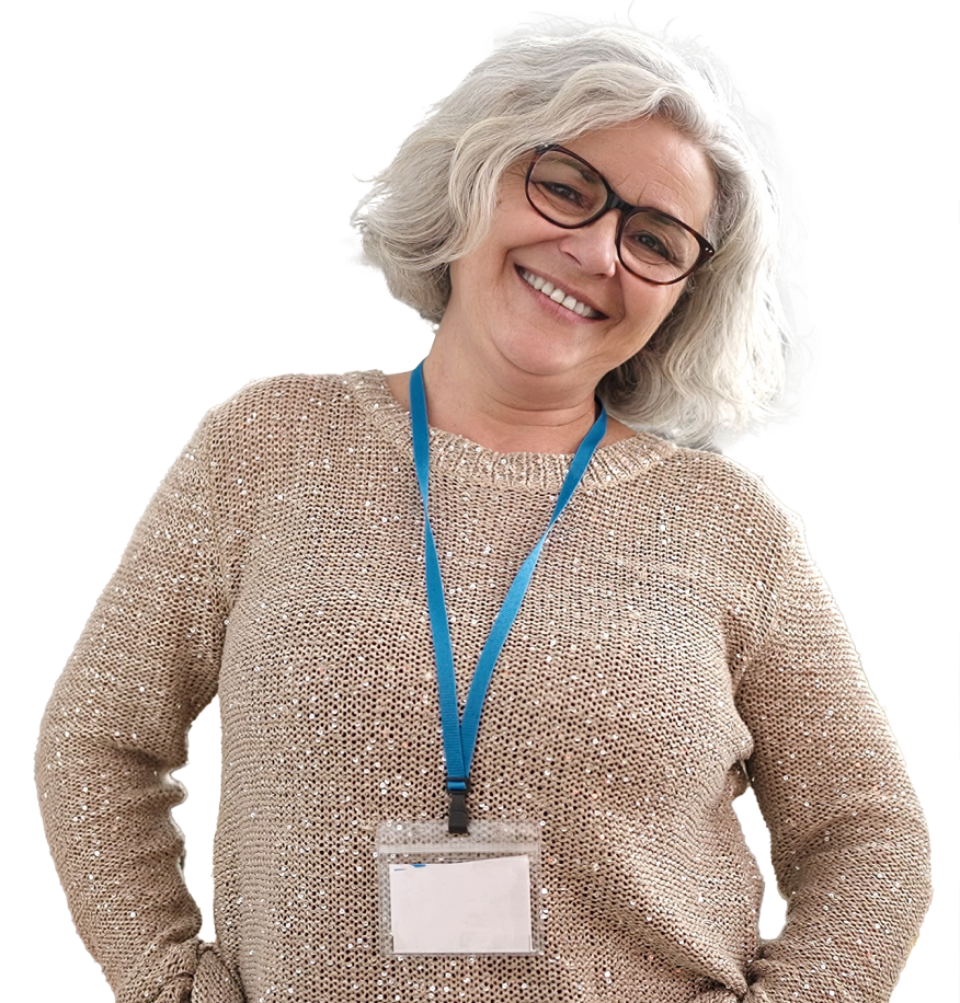 A white woman with white hair and dark glasses posed with her head tilted to one side while smiling at the camera with her hands in her front pockets. She is wearing a beige sweater, jeans, and a conference badge and lanyard.